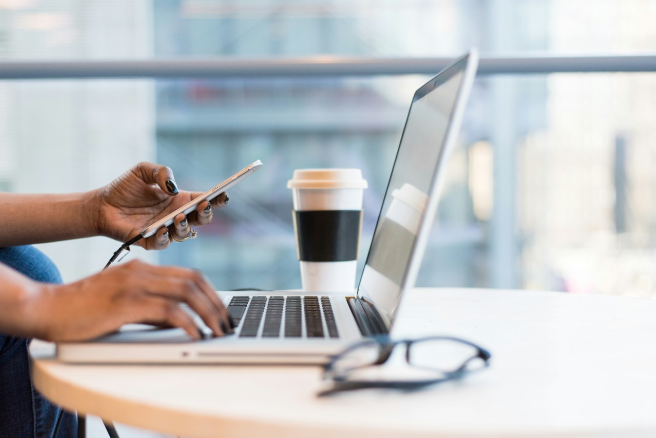 Person multitasking at a desk, holding a smartphone while typing on a laptop, with a to-go coffee cup and glasses nearby.